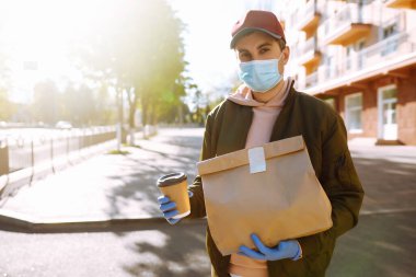 Koruyucu maskeli ve tıbbi eldivenli kurye paket yiyecek ve kahve getiriyor. Karantina şehrinde teslimat servisi. Covid-19. Evde kal..