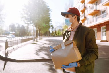 Koruyucu maskeli ve tıbbi eldivenli kurye, içinde yiyecek olan kağıt paketler getiriyor. Karantina şehrinde teslimat servisi. Covid 19.