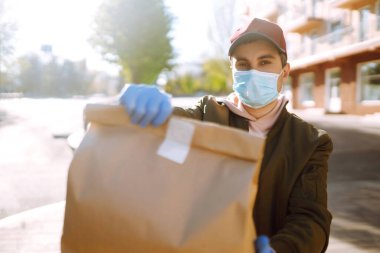 Koruyucu maskeli ve tıbbi eldivenli kurye, içinde yiyecek olan kağıt paketler getiriyor. Karantina şehrinde teslimat servisi. Covid 19.