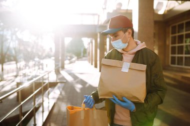 Koruyucu maskeli ve tıbbi eldivenli kurye, içinde yiyecek olan kağıt paketler getiriyor. Karantina şehrinde teslimat servisi. Covid 19.