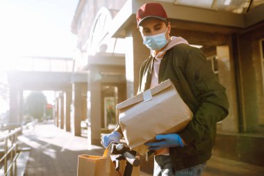 Koruyucu maskeli bir kurye ve scooterlı tıbbi eldivenler içinde yiyecek olan kağıt paketler getiriyor. Karantina, hastalık salgını, koronavirüs covid-19 salgını altında teslimat hizmeti.