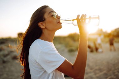 Gün batımında sahilde bira içen güzel kız. Genç bir kadın plaj tatilinde eğleniyor. Yaz, rahatla ve yaşam tarzı kavramı.