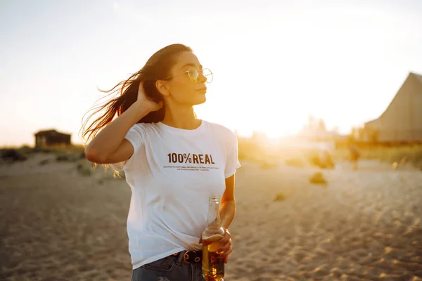 Pretty girl drinking beer at the beach at sunset. Young woman enjoying on beach holiday. Summer, relax and lifestyle concept.