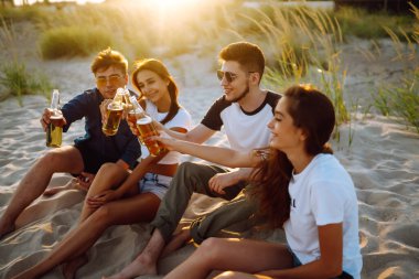 Bir grup genç arkadaş gün batımında sahilde oturup sohbet ediyor ve bira içiyor. Yaz tatili, tatil, rahatlama ve yaşam tarzı kutsallığı.