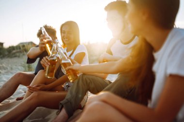 Bir grup genç arkadaş gün batımında sahilde oturup sohbet ediyor ve bira içiyor. Yaz tatili, tatil, rahatlama ve yaşam tarzı kutsallığı.