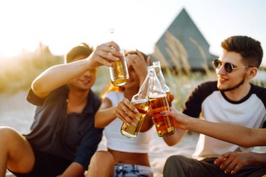 Bir grup genç arkadaş gün batımında sahilde oturup sohbet ediyor ve bira içiyor. Yaz tatili, tatil, rahatlama ve yaşam tarzı kutsallığı.