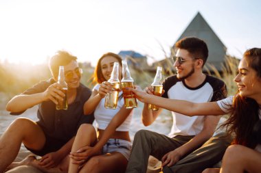 Bir grup genç arkadaş gün batımında sahilde oturup sohbet ediyor ve bira içiyor. Yaz tatili, tatil, rahatlama ve yaşam tarzı kutsallığı.