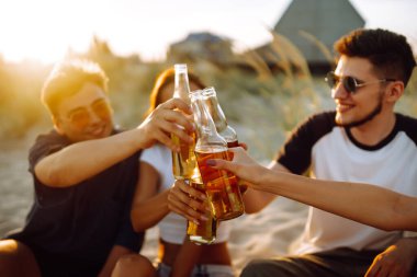 Bir grup genç arkadaş gün batımında sahilde oturup sohbet ediyor ve bira içiyor. Yaz tatili, tatil, rahatlama ve yaşam tarzı kutsallığı.