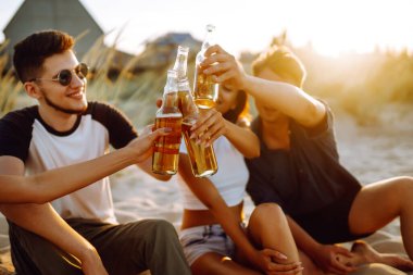Bir grup genç arkadaş gün batımında sahilde oturup sohbet ediyor ve bira içiyor. Yaz tatili, tatil, rahatlama ve yaşam tarzı kutsallığı.