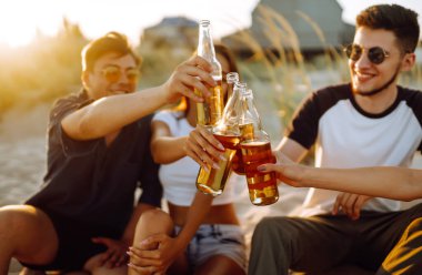 Bir grup genç arkadaş gün batımında sahilde oturup sohbet ediyor ve bira içiyor. Yaz tatili, tatil, rahatlama ve yaşam tarzı kutsallığı.