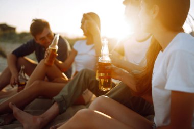 Bir grup genç arkadaş gün batımında sahilde oturup sohbet ediyor ve bira içiyor. Yaz tatili, tatil, rahatlama ve yaşam tarzı kutsallığı.