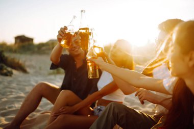 Bir grup genç arkadaş gün batımında sahilde oturup sohbet ediyor ve bira içiyor. Yaz tatili, tatil, rahatlama ve yaşam tarzı kutsallığı.