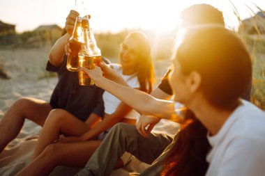 Bir grup genç arkadaş gün batımında sahilde oturup sohbet ediyor ve bira içiyor. Yaz tatili, tatil, rahatlama ve yaşam tarzı kutsallığı.