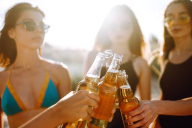 İnce kızlar gün batımında sahilde bira içip tezahürat yaparlar. Bikinili dört kız plaj tatilinde eğleniyor. Yaz tatili, tatil, rahatlama ve yaşam tarzı konsepti.