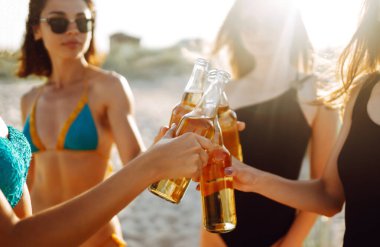İnce kızlar gün batımında sahilde bira içip tezahürat yaparlar. Bikinili dört kız plaj tatilinde eğleniyor. Yaz tatili, tatil, rahatlama ve yaşam tarzı konsepti.