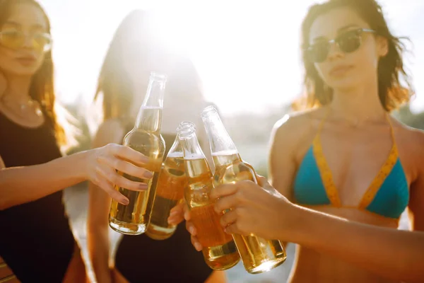 Slender girls cheers and drink beers on the beach at sunset. Young four girl in bikini enjoying on beach holiday.  Summer holidays, vacation, relax and lifestyle concept.
