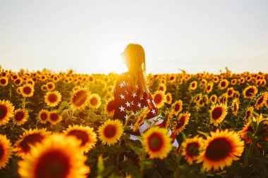 4 Temmuz. Ayçiçeği tarlasında Amerikan bayrağı taşıyan hoş bir kız. Özgürlük. Gün batımı ışığı. Bağımsızlık Günü. Vatansever kavram.