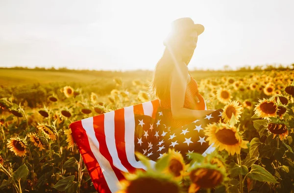 7月4日戴着帽子 戴着美国国旗 站在葵花田里的漂亮姑娘 自由日夕阳西下独立日 爱国观念 — 图库照片