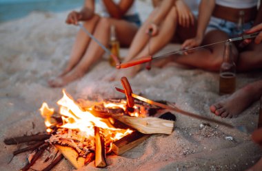 Şenlik ateşinde kızarmış sosis. Bir grup genç arkadaş sahilde oturup sosis kızartıyorlar. Yaz tatili, tatil, rahatlama ve yaşam tarzı kutsanması. Kamp zamanı..
