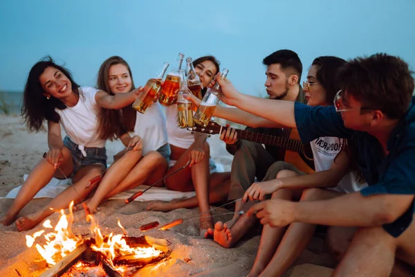 Group of friends near a campfire on the beach cheers and drinking beer at night. Summer holidays, vacation, relax and lifestyle concept.