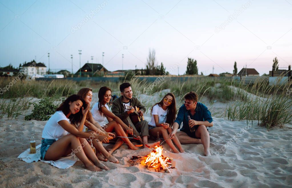 Group of young friends sitting on beach and fry sausages. One man is playing guitar. Summer holidays, vacation, relax and lifestyle consept. Camping time.