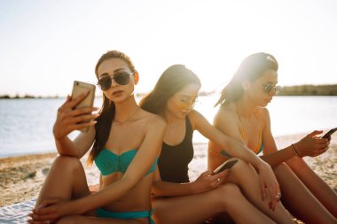Sahilde cep telefonu kullanan üç güzel kız. Kızlar kumsalda güneşleniyor. Genç kızlar telefonla selfie çekiyorlar. Yaz tatilinin tadını çıkar.