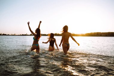Üç genç kız gün batımında denizde yüzmek için koşar. Yaz tatili, tatil, rahatlama ve yaşam tarzı konsepti.