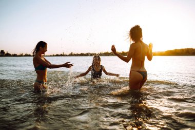 Denize su sıçratan üç genç kız. Mutlu arkadaşlar gün batımında sahilde eğleniyorlar. Yaz tatili, tatil, rahatlama ve yaşam tarzı konsepti.