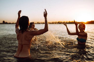 Denize su sıçratan üç genç kız. Mutlu arkadaşlar gün batımında sahilde eğleniyorlar. Yaz tatili, tatil, rahatlama ve yaşam tarzı konsepti.