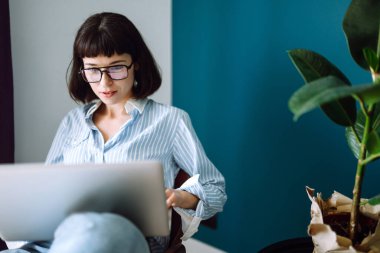 Dizüstü bilgisayarla çalışan genç bir kadın evdeki modern oturma odasında oturuyor. Sosyal ağda arkadaşlarıyla sohbet eden, internetten alışveriş yapan, e-posta yazan kız. Teknoloji, serbest meslek ve iş kavramı.