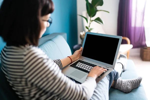 Young girl sitting on a sofa with a laptop at home. Black screen on laptop. Technology, freelance and work concept.