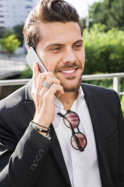 genç yakışıklı gülümseyen businessmanin siyah takım elbise gömlek, bilezik ve Aksesuar ayakta dışında bir kafede ve cep telefonu üzerinde konuşurken elleri ile.