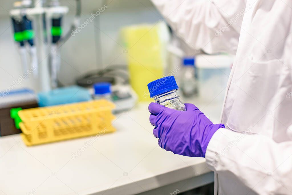 Female hand in blue gloves in bathrobe in laboratory holds bottle