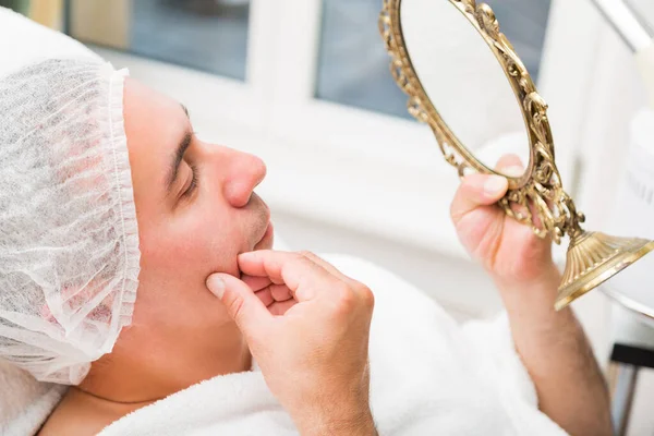 Мужчина рассматривает лицо в зеркале, лежащем в салоне красоты — стоковое фото
