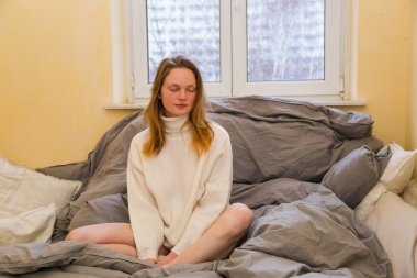 Sarışın, uzun saçlı. Yatakta süveter giyiyor.