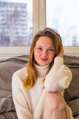 Kazaklı kız pencerenin yanında dizlerine sarılıyor.