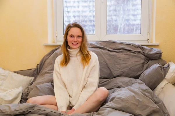 Sarışın, uzun saçlı. Yatakta süveter giyiyor.