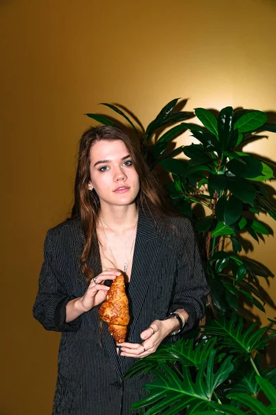 Menina com croissant fica perto de planta no estúdio — Fotografia de Stock
