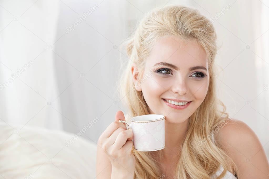 Beautiful young smiling woman sitting on white bed , drinking a cup of coffee