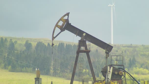 Bomba de pozo de aceite y planta de energía eólica en el fondo con cielo gris — Vídeos de Stock