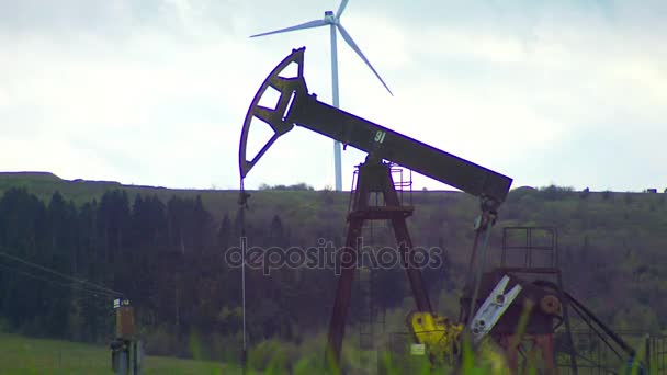 Pozo de aceite con toma de bomba y planta de energía eólica en la montaña en el fondo — Vídeo de stock
