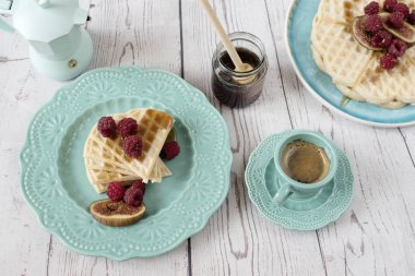 Ahududu ve incir, gözleme şeklinde yumuşak Belçika kalp turkuaz mavi tabakta bal ile kaplı. Bir kahve ve kahve makinesi. Rustik ahşap arka, üstten görünüm. Tatil sabah evde
