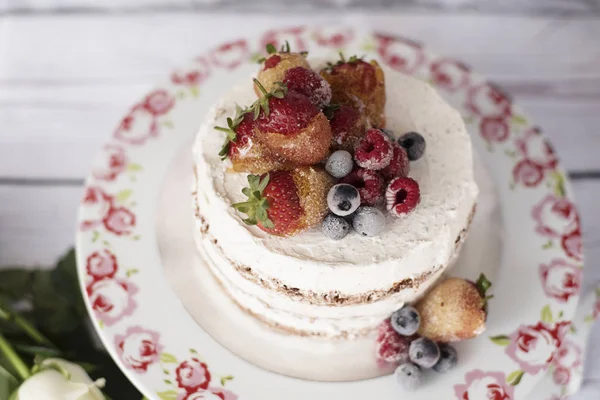 Bolo nu com frutas caramelizadas - morangos, mirtilos, framboesas. Bolo de creme de esponja em planalto alto floral, bandeja. Rosas brancas, fundo rústico — Fotografia de Stock