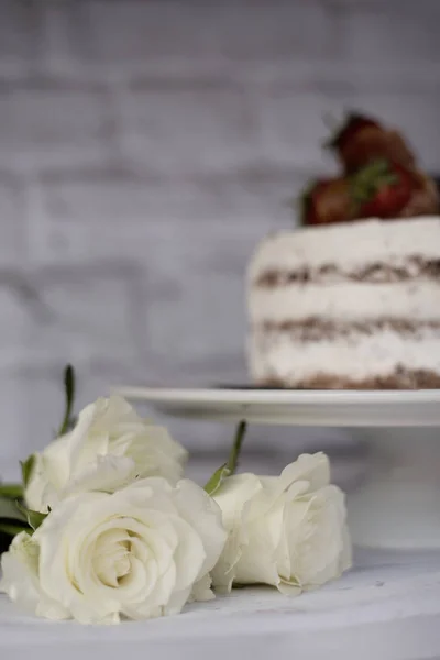 Três rosas brancas. Bolo de chocolate nu desfocado com creme atrás. Um fundo rústico brilhante. Foco seletivo. Imagem vertical — Fotografia de Stock