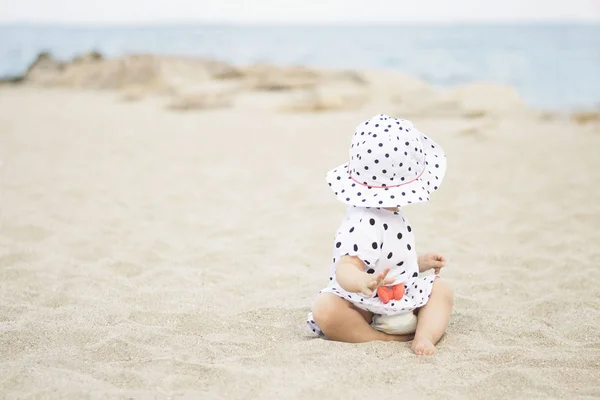 Linda niña con un vestido y un sombrero en lunares sentados en la arena en la orilla del mar y mira hacia el mar — Foto de Stock