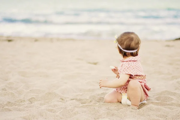Schattige babymeisje met een jurk op polka dots zittend op het zand op de kust en kijkt uit naar de zee — Stockfoto