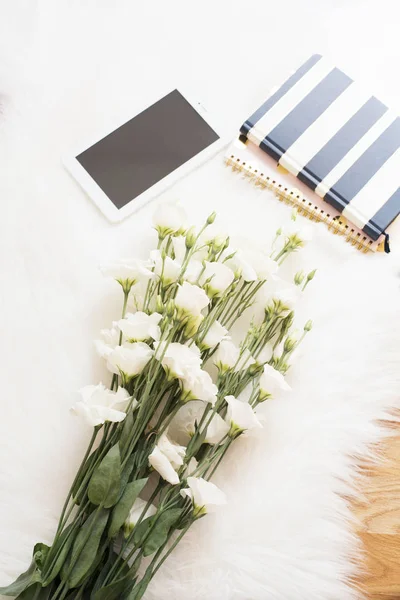 Un gran ramo de flores blancas, cuadernos y tabletas en el suelo sobre una alfombra de piel blanca. Acogedor, moda cómoda feminidad hogar lugar de trabajo. Estilo laico plano. Vista superior, imagen vertical —  Fotos de Stock