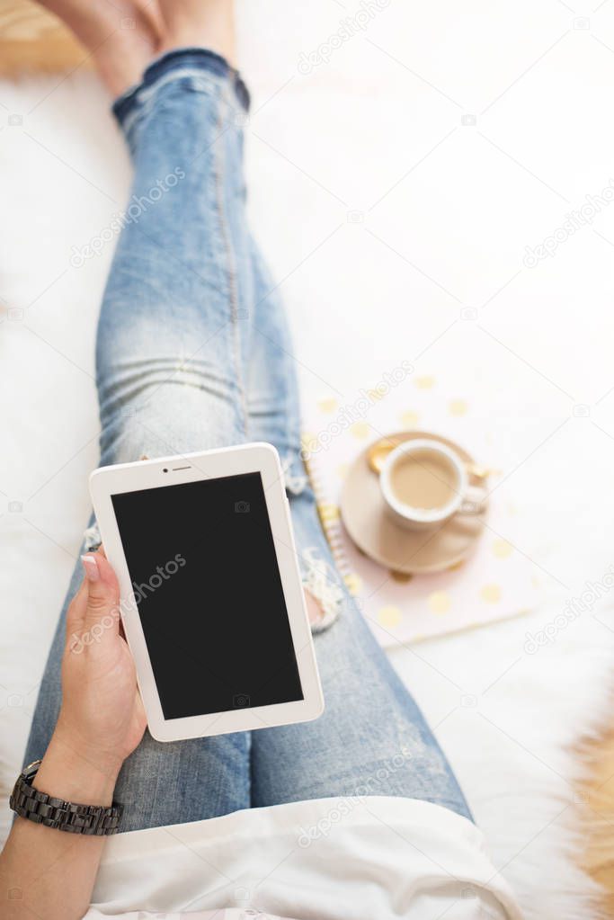 A young woman wearing distressed jeans sitting on wood floor on a white fur carpet at home and holds a tablet in his hand. Around a cup of coffee, notebooks. Gold bright feminine theme