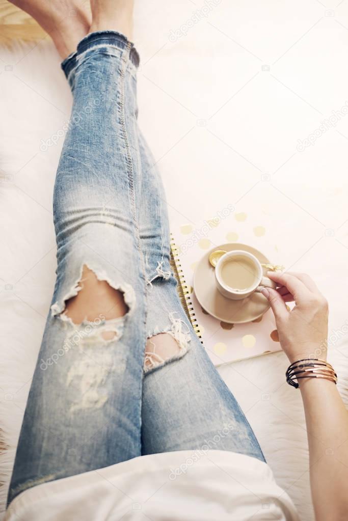 A young woman wearing distressed jeans sitting on wood floor on a white fur carpet at home and holds a cup of coffee in his hand. Gold bright feminine theme