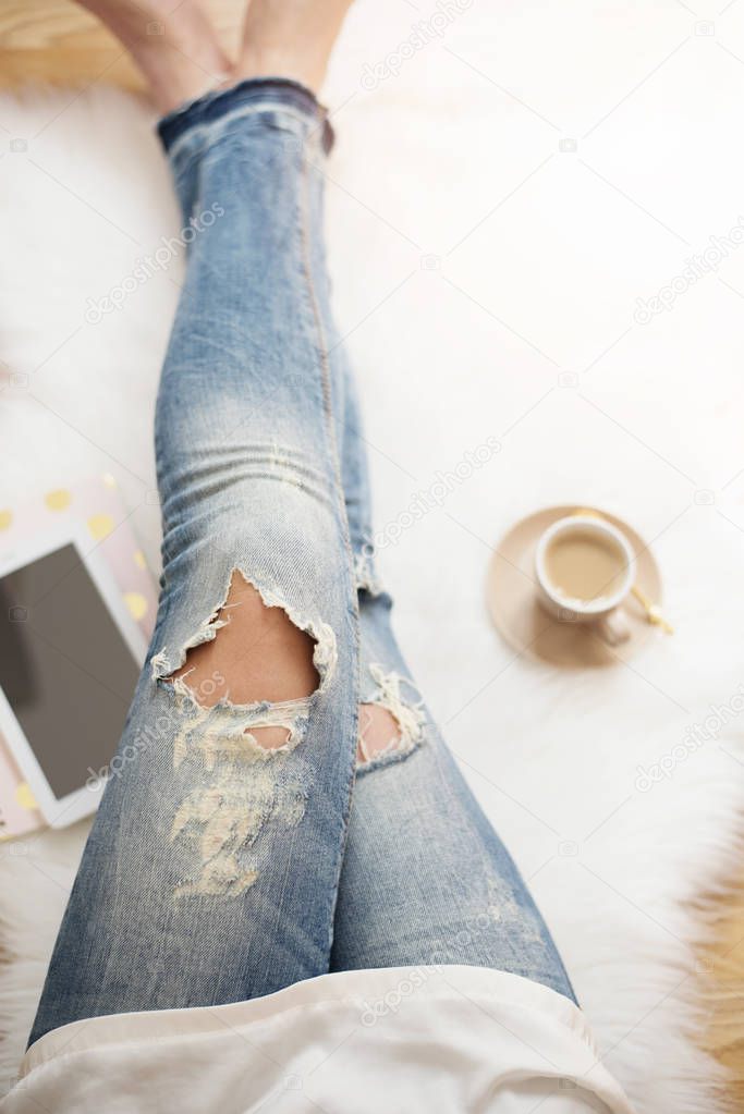 A young woman wearing distressed jeans sitting on wood floor on a white fur carpet at home. Around a cup of coffee, tablet and notebooks. Gold bright feminine theme